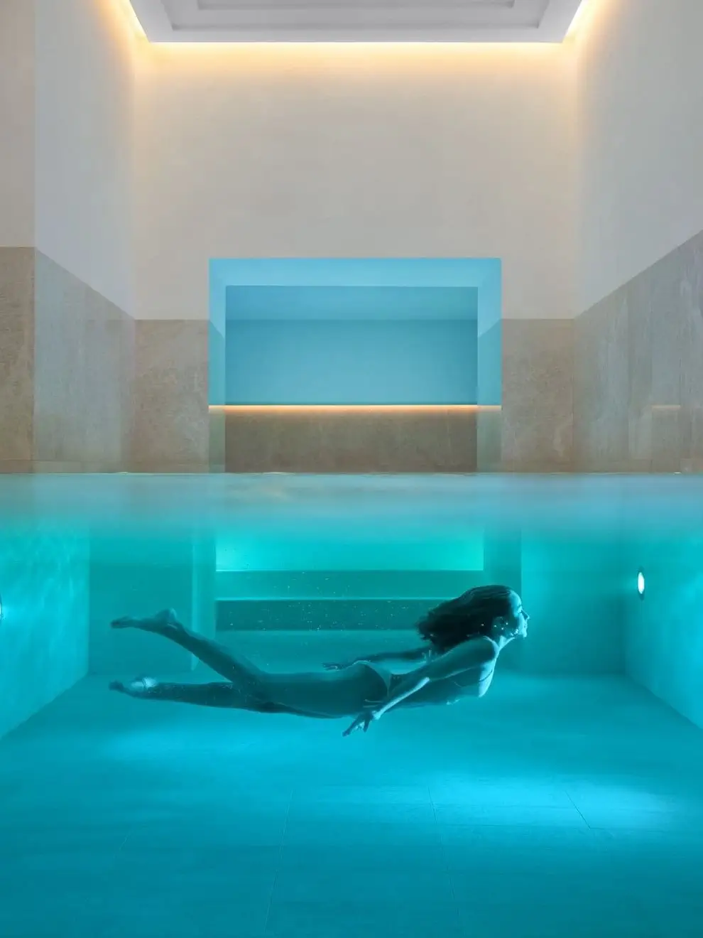 Eine Person im Badeanzug schwimmt unter Wasser in einem modernen Hallenbad mit blauer Beleuchtung und beigen Wänden.